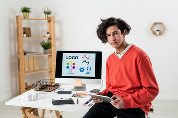 Young serious freelance designer with touchpad sitting on desk at home office