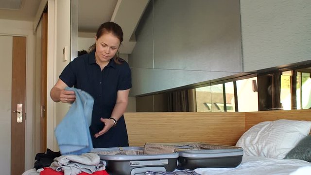 Happy girl preparing her suitcase for a travel, she is packing her clothes in the bedroom, tourism and vacations concept. Folding clothes into a suitcase, a woman smiles as she prepares for a journey.