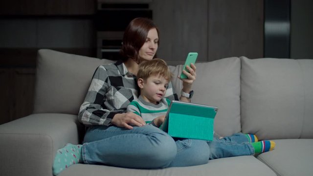 Mother with smartphone hugging son with tablet computer sitting on cozy sofa. Family using gadgets phubbing each other. 