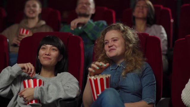 Tracking left medium shot of laughing young people enjoying watching comedy movie sitting on comfortable seats in cinema. Female friends eating popcorn and discussing funny episodes