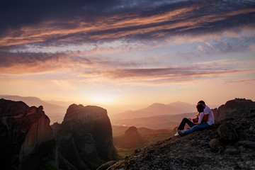 Stunning Nature Landscape. Romantic couple in love sitting together at sunset on the Mountain, silhouettes of young man and woman during sunset over the Alpine mountains valley with Colorful sky