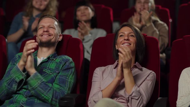 Tracking left medium shot of happy audience applauding after watching movie in cinema. Middle aged couple exchanging opinions about film