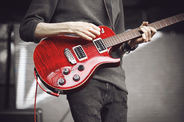 A guitarist in a grey sweater and dark jeans with a bright red electric guitar plays a powerful...