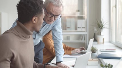 Waist-up shot of senior Caucasian male manager walking up to young Afro-American trainee, looking at his work on laptop, talking, listening, them giving him feedback and advice