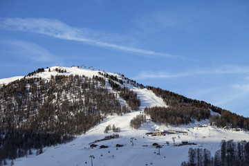 Snowy ski slope with ski-lifts, traces and forest in high winter mountains at sunny morning