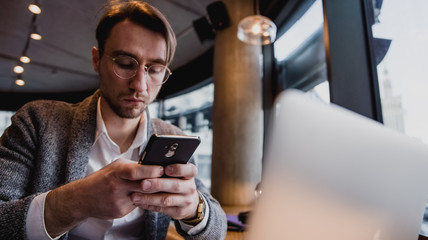 Businessman holding smartphone in hands, touching screen, texting message, chatting in social networks, meeting website, searching internet, sending sms, using text messenger. Laptop on the table, 