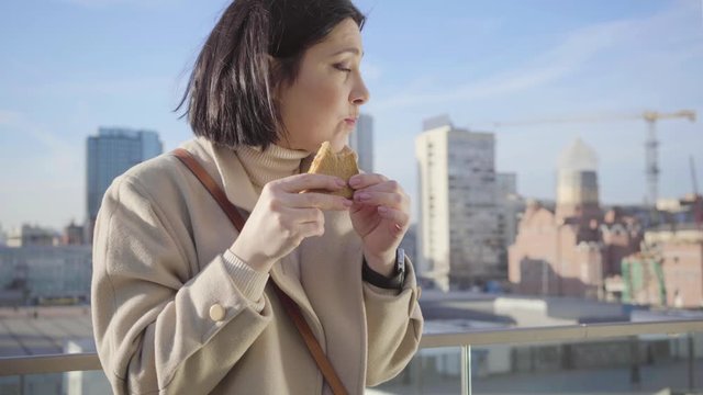 Portrait of young brunette Caucasian woman eating hungrily sandwich. Successful businesslady having break on the city street. Overworking, body weight problems, junk food.