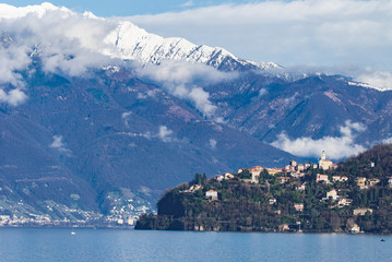 vue sur le Lago Maggiore d'un village sur une colline au soleil et des montagnes enneigées et pointues au loin