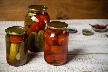 Three cans of assorted fermented tomatoes and cucumbers. In the background, saucers with seasonings: cloves, sweet and red, hot pepper. Stand on a textured, white background, horizontal arrangement.