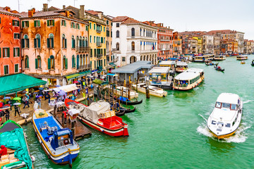 Fototapeta na wymiar Water taxis, vaporettos, boats, gondolas sailing and docked on Grand Canal along wooden mooring poles and colorful Venetian architecture buildings. People/ tourists in Venice city, Italy on rainy day.
