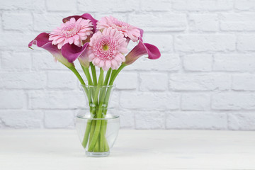Bouquet of pink calla lilies and Gerber daisies in a clear vase isolated on a white brick background