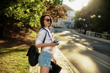 The girl in the black hat and sun glasses with photo camera on a road in an old european town.