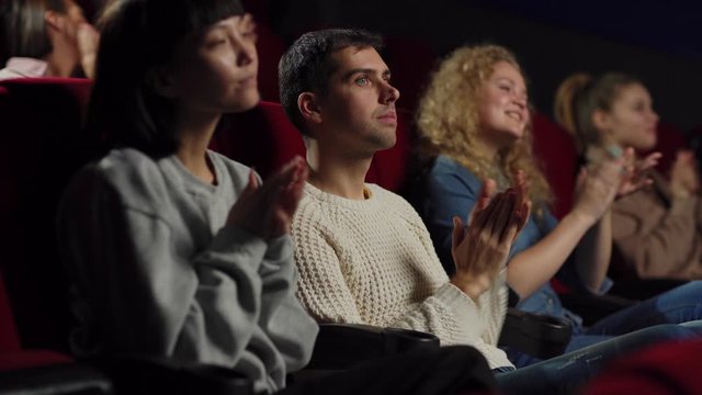 Side view medium shot of satisfied audience applauding after watching movie in cinema, focus on Caucasian young man