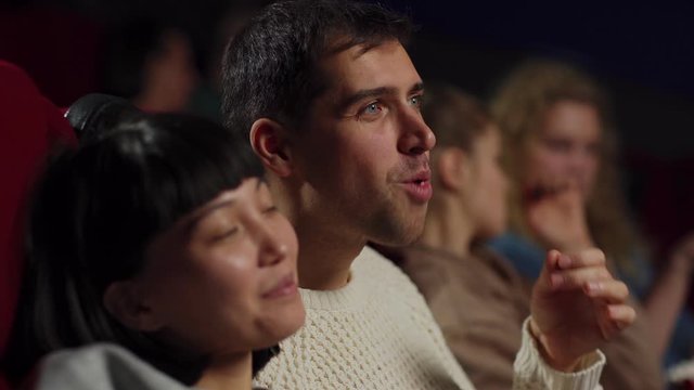 Side view closeup of cheerful diverse young couple of Asian woman and Caucasian man eating popcorn, talking and smiling while watching movie in cinema