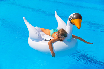 Pretty tanned young girl in swimsuit playing and sunbathing on inflatable swan in blue swimming pool in family vacation. Happy child relaxing on holidays. Summertime. Rest. Resort. Active