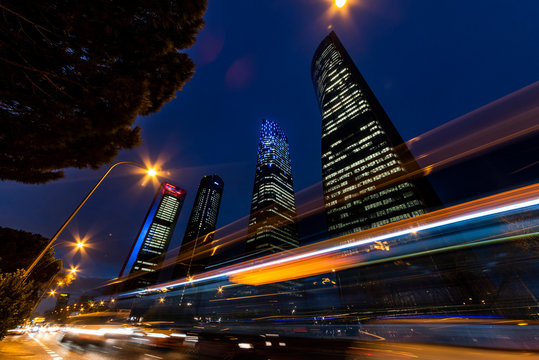 Vista de Cuatro Torres Business Area en Madrid al anochecer con las luces de las ventanas encendidas y las estelas de los coches pasando.