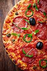 Pizza with salami, jalapeno pepper, black olives, capers and fresh herbs. Home made food. Concept for a tasty and hearty meal. Dark wooden background. Top view.