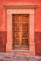 A pic of a patterned varnished wooden door with a cement frame and a reddish wall