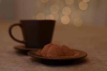 Brown tea steam with soft lights hovering over the cup and chocolate candy truffles, sprinkled with cocoa, on beige.