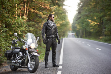 Obraz premium Young bearded tall athletic motorcyclist in black leather outfit and dark sunglasses standing at shiny modern powerful motorbike, on blurred background of hilly asphalt road and green trees.