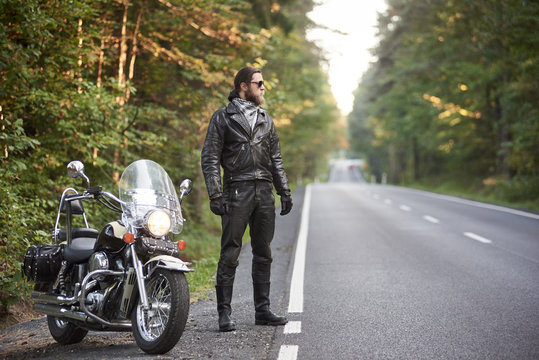 Young bearded tall athletic motorcyclist in black leather outfit and dark sunglasses standing at shiny modern powerful motorbike, on blurred background of hilly asphalt road and green trees.