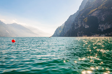 Scenic view against bright sun rays above the rocks on orange buoy floating in beautiful Garda lake in Italy surrounded by high dolomite mountains and crystal clear blue water of the lake
