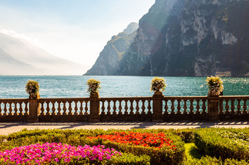 Garda lake promenade with colorful flowerbeds with growing and blooming plants, classic stone fence built on the edge with flowerpots with blooming flowers. Garda lake and mountains on background