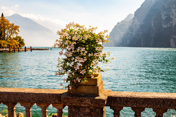 Beautiful Garda lake promenade with classic stone fence railings built on the edge with flowerpots with blooming white flowers. Garda lake surrounded by the high dolomite mountains on the background