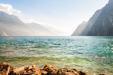 Rocky stones lying on the shore of beautiful Garda lake in Lombardy, Italy surrounded by high dolomite mountains. Sunbeams penetrating from above the rocks and warming misty fog above the water