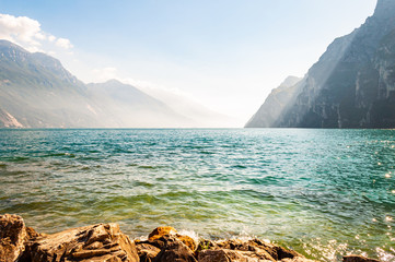 Rocky stones lying on the shore of beautiful Garda lake in Lombardy, Italy surrounded by high dolomite mountains. Sunbeams penetrating from above the rocks and warming misty fog above the water
