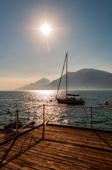 Sailing yacht floating near the pier on misty Garda lake with high dolomite mountains with sun shining above in the sky on the background. Northern Italy, Lombardy