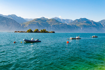 Motor boats on water, locked to parking lots floating near the shore of Garda lake with small island overgrown with trees from behind and high dolomite mountains on the background. Lombardy, Italy