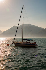 Sailing yacht floating near shore of misty Garda lake with high dolomite mountains with sun shining above in the sky on the background. Northern Italy, Lombardy