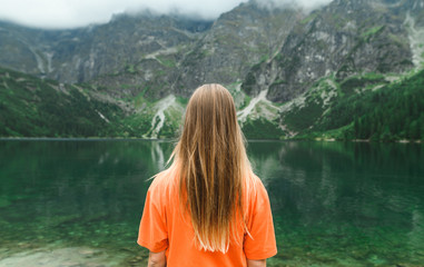 Back of young hiker girl in orange football stands on the background of lake in mountains and looks...