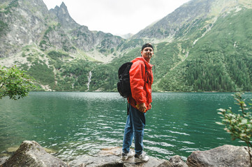 Portrait of happy young male tourist in red raincoat stands on background of lake and green mountains with coniferous trees, looks into camera and smiles. Morskie Oko, Tatra Mountains
