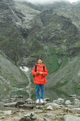 Full-length vertical portrait of a funny young hiker man in a red jacket stands with a smartphone in hand against the background of mountains and lakes, looks into the camera and smiles.Man and nature
