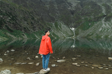 Full length photo of a man standing on a rock against a lake background at the top of a mountain, looking to the side, wearing a red jacket and cap.Hiker guy hiking in the mountains enjoys the scenery