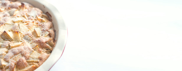 Part of homemade baked apple cake sprinkled with icing sugar in a white round ceramic baking dish on a white background, top view, close-up, banner.