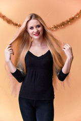 Fototapeta premium Portrait of a cute girl in a black T-shirt with long beautiful hair and great makeup. Concept of a young blonde woman with New Year's decor. Smiling, showing emotions on a pink background.