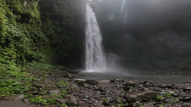 A big waterfall crashing down on big, black rocks in the jungle of Bali, camera dolly shot forward.