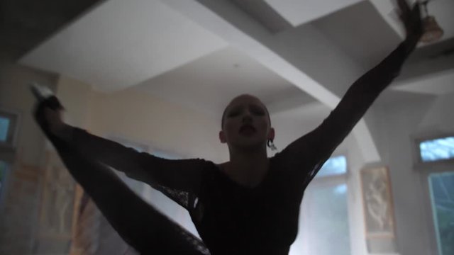 A mysterious young woman with shaved head is holding a smoke grenade and dancing dramatically back and forth while spreading the smoke around the room.