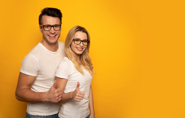 Smart cool people. Close-up photo of man and woman in casual outfits and glasses, who are looking in the camera with broad smiles and showing thumbs up.