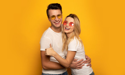 Funky and funny. A charming hipster couple in multicolored sunglasses is hugging each other and laughing, while the girl is showing thumbs up.