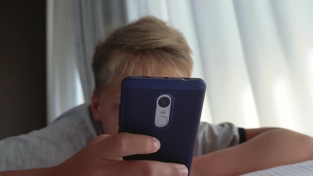 Closeup view of white cute kid laying in bed and using modern smartphone. Real time wide angle 4k video footage.
