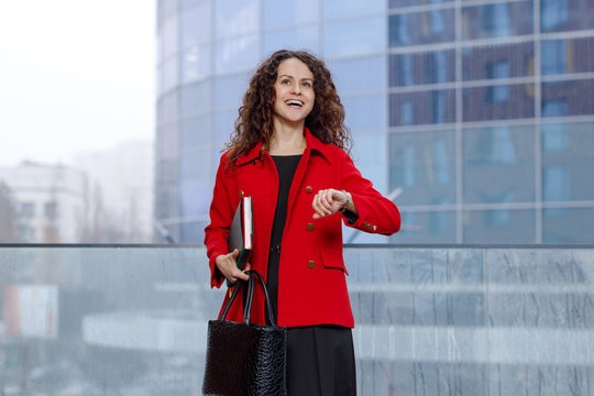 Horizontal portrait of a cheerful business woman, in red suit, coming on a job, look at the time, holding in hands agend and elegant bag.
