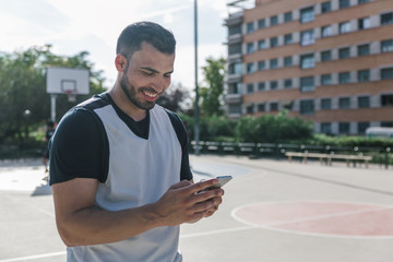 Attractive sportsman man looks at his mobile phone while taking a break in the middle of an urban court