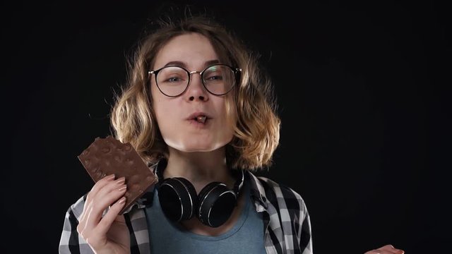 Close up brunette young woman with glasses and headphones posing playfully eat, chewing chocolate bar with nuts isolated over black background in studio. People sincere emotions lifestyle concept