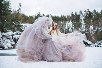 Blonde woman in waving dress in winter outdoors. Fabric flying and fluttering. Fashion model in long waving gown dancing on frozen lake covered with snow. Winter and snow queen