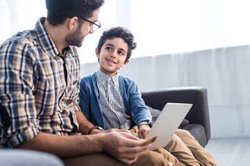 Jewish father and smiling son using laptop in apartment