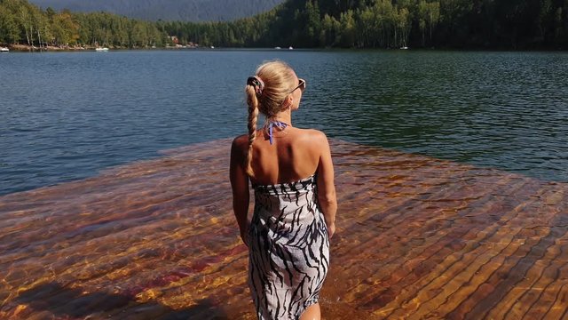 Woman walk on water on pier in sunglasses and a boho silk shawl. Girl rest on a flood wood underwater dock. The pavement is covered with water in the lake. In the background are mountain and a forest.
